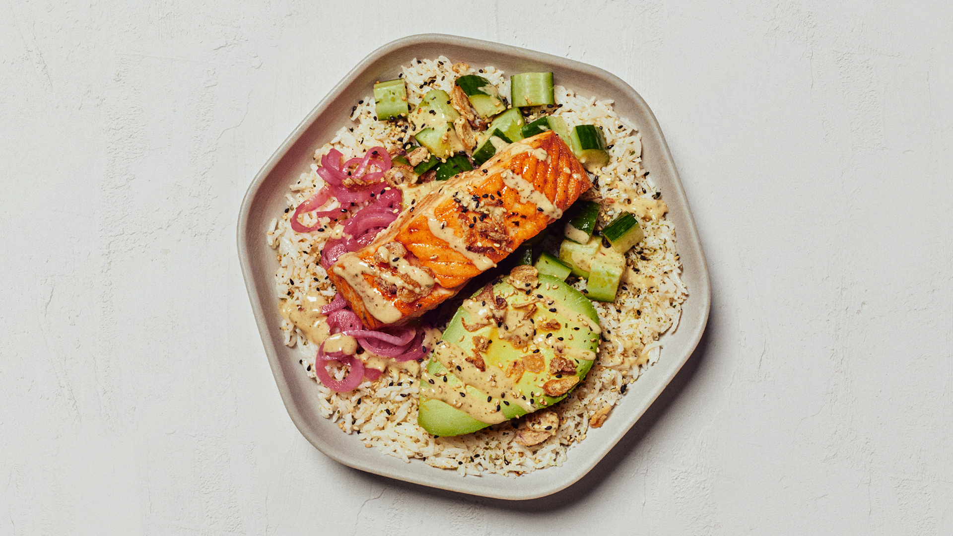 A hexagonal plate of a filet of salmon and an avocado, served on rice and drizzled with sauce