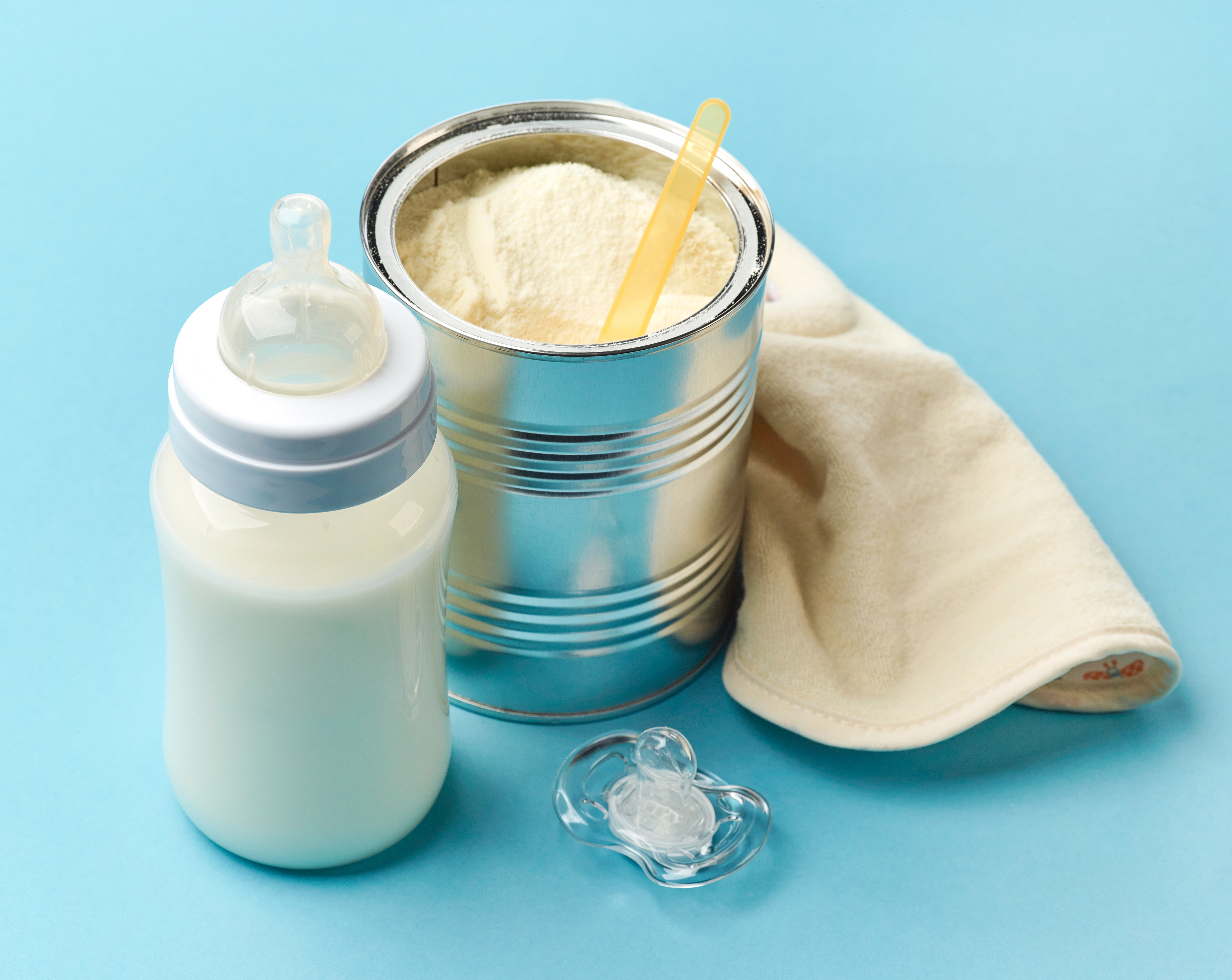 Open can of baby formula with a scoop inside of it next to a bottle filled with white liquid and a pacifier.