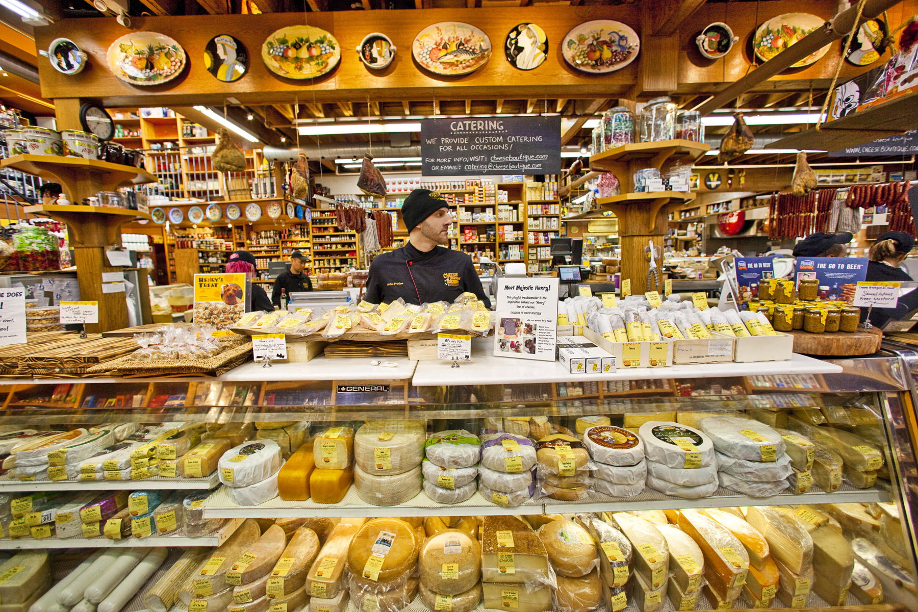 A worker looks away from the camera behind a deli case filled with cheese, with more cheese all around.