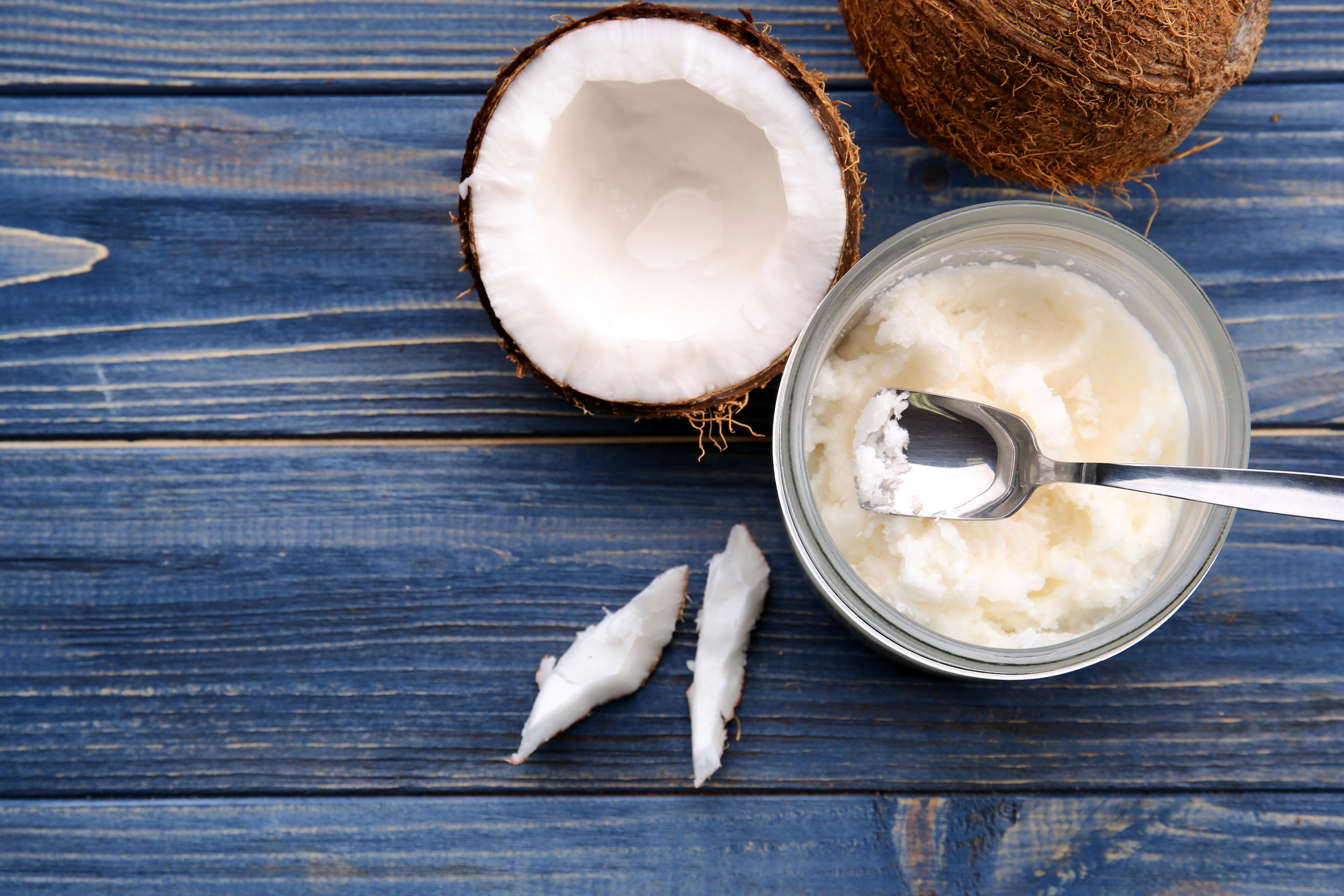 Overhead view of a half of an open coconut and jar of coconut oil with a spoon in it, both on a table top.