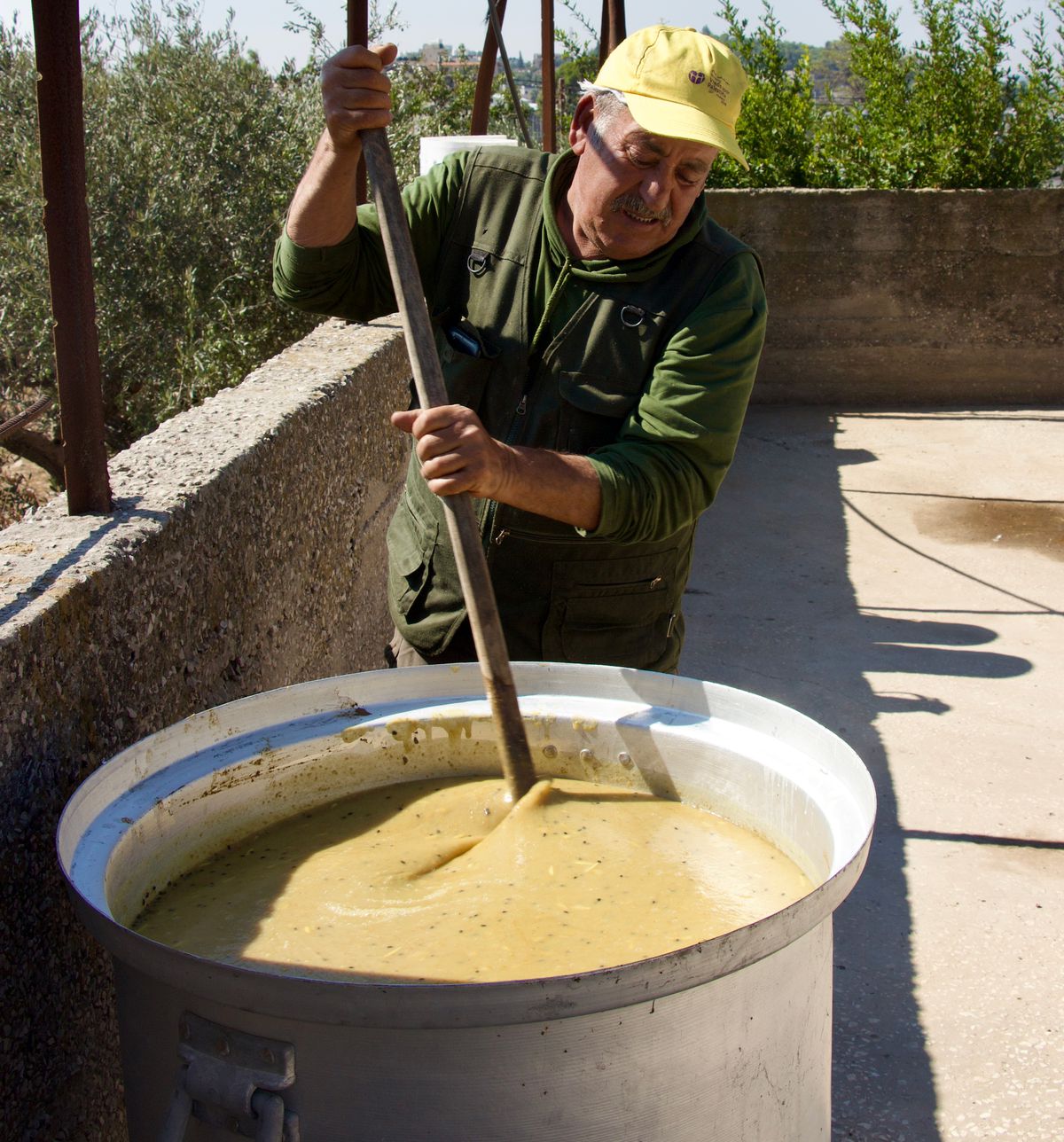 A worker stands beside a large pot stirring a yellow substance with a long pole.