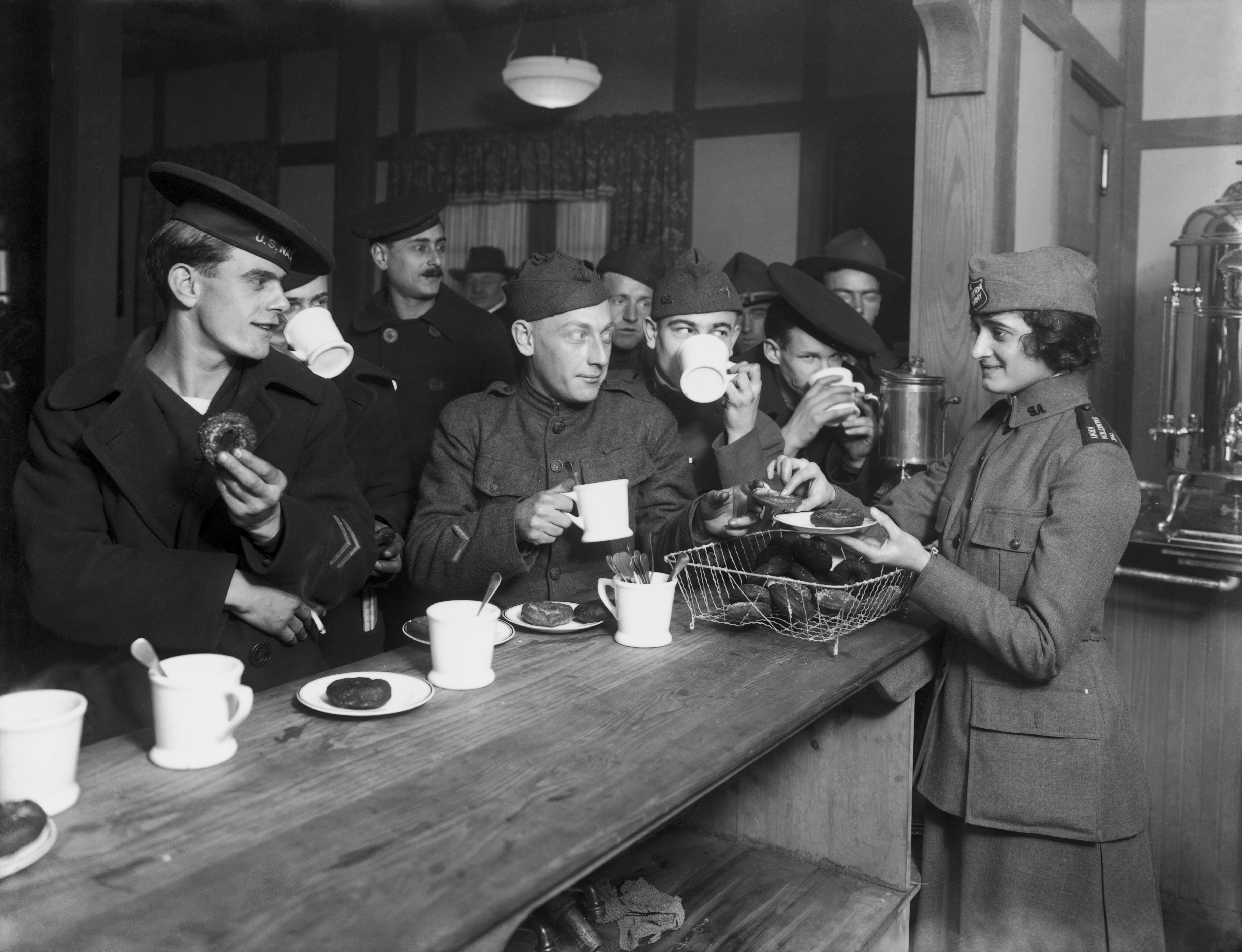 A group of soldiers congregates at a bar to sip coffee and eat doughnuts as a woman working with the Salvation Army distributes the o-shaped confections to the crowd of hungry-looking men.
