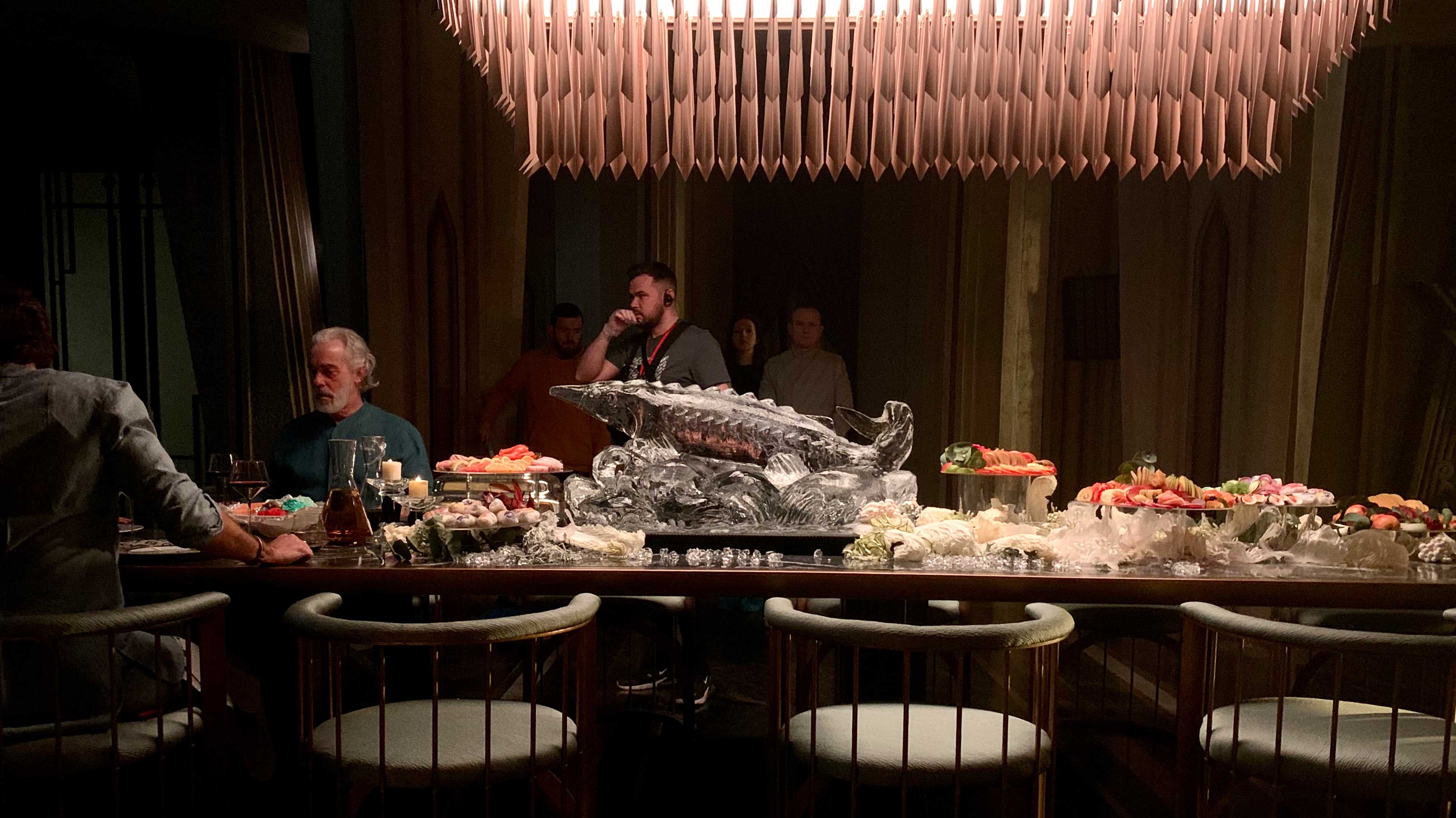 An elaborate tablescape under a chandelier sits on a film set, with two actors seated and a man directing behind him.