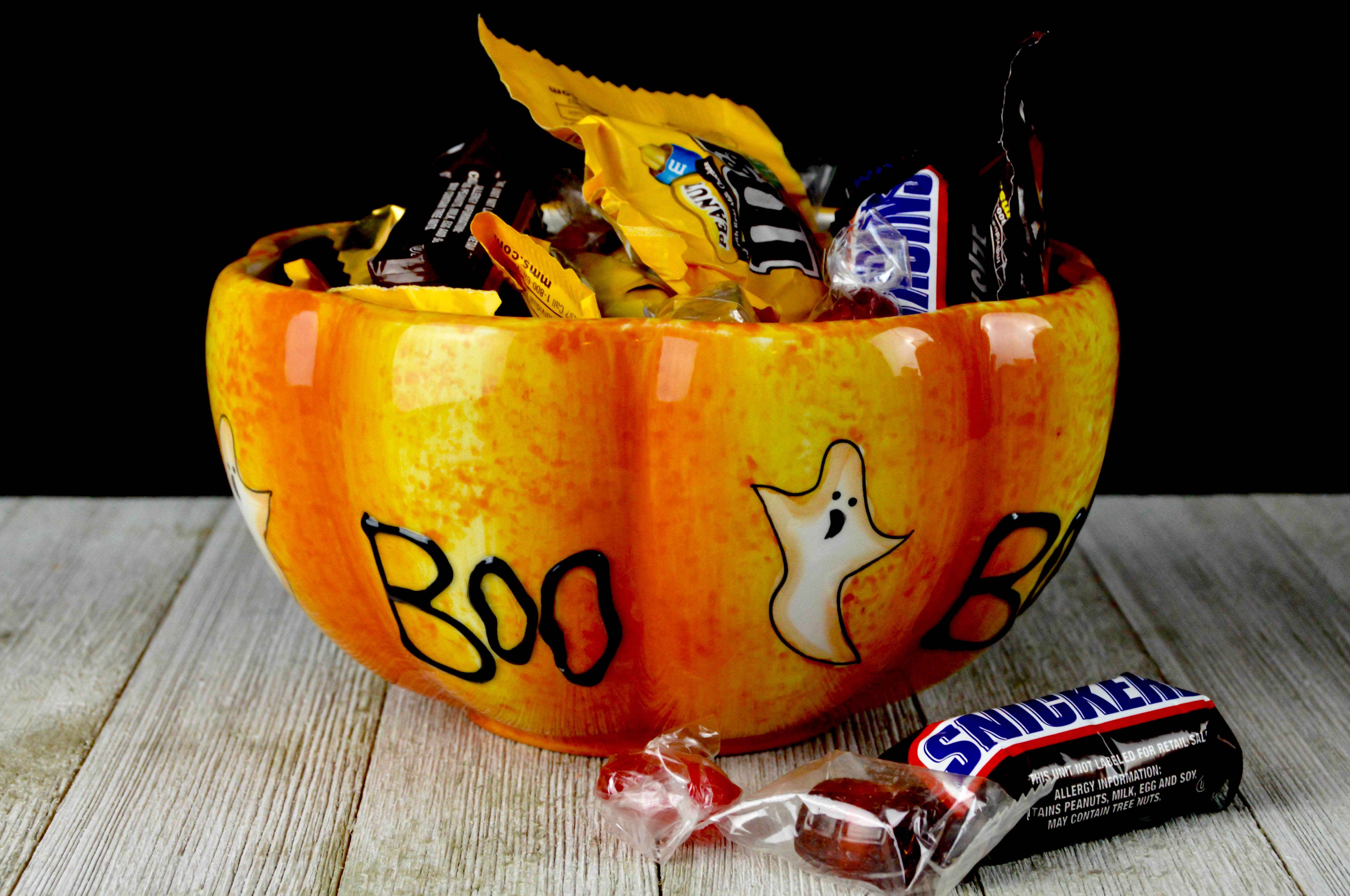 An orange ceramic bowl, shaped like the bottom half of a pumpkin and painted with ghosts and the word “boo,” overflows with name-brand candy like Snickers and peanut M&amp;Ms. 
