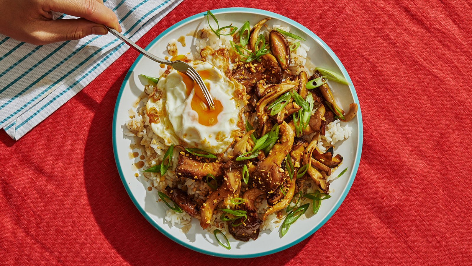 A hand holding a fork breaks a fried egg yolk that sits atop a bowl of mushroom adobo and rice, garnished with scallions.